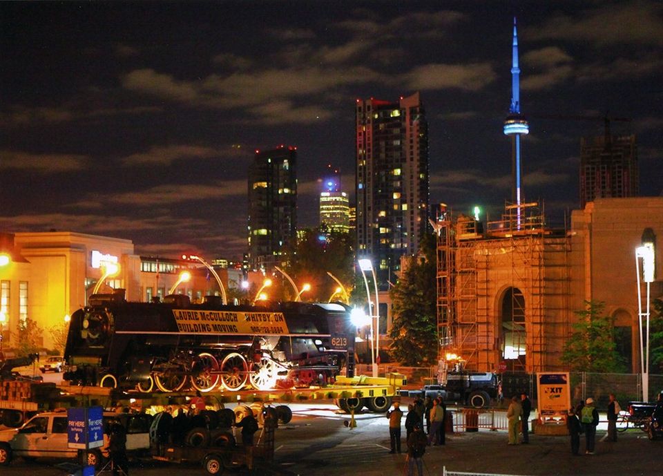 Nighttime photograph of the engine on top of a trailer, ready to move. The CN tower, all lit up, can be seen in the background.