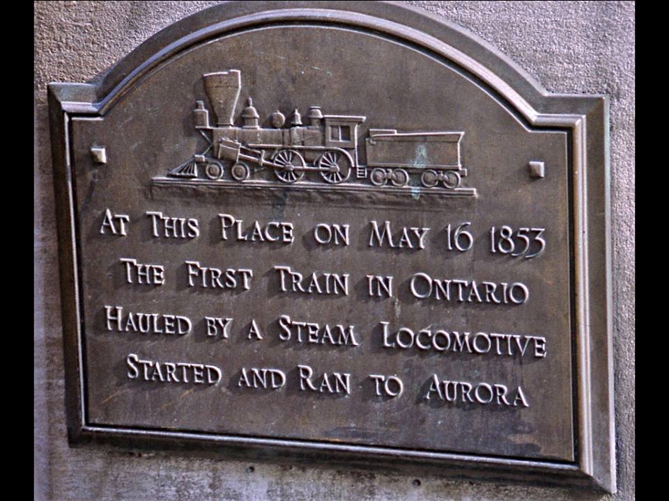 Close-up shot of a metal plaque hung on a stone wall reads "at this place on May 16, 1853 the first train in Ontario hauled by a steam locomotive started and ran to Aurora."
