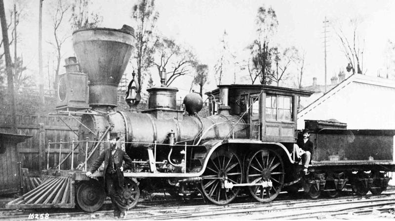 Black and white photograph of an early steam locomotive. Two men pose in front of it.