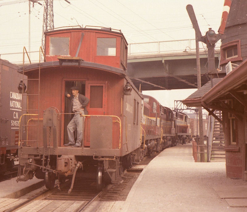 A man leans against an open caboose doorway, at the end of a train parked at Don Station. To the left, the edge of the station can be seen, notably it's two semaphore signal arms.