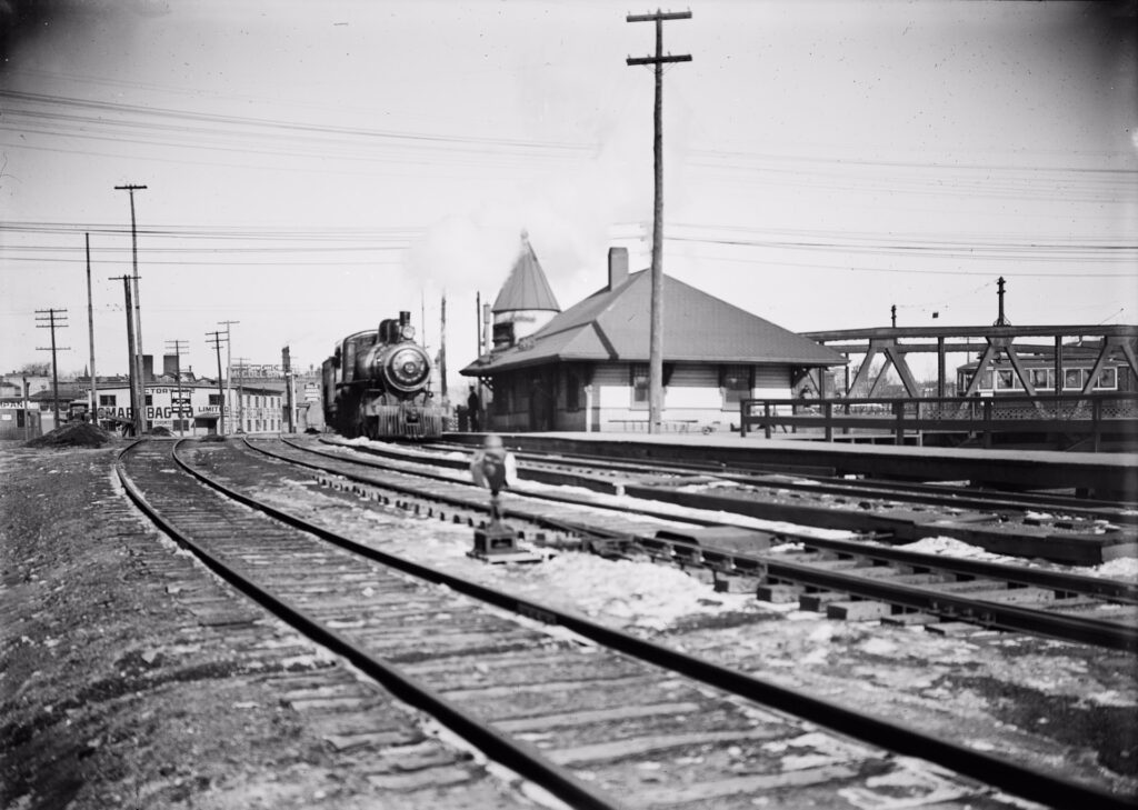 Black and white historical photo of Don Station taken at track level with a view of the station and a steam locomotive stopped at the platform.