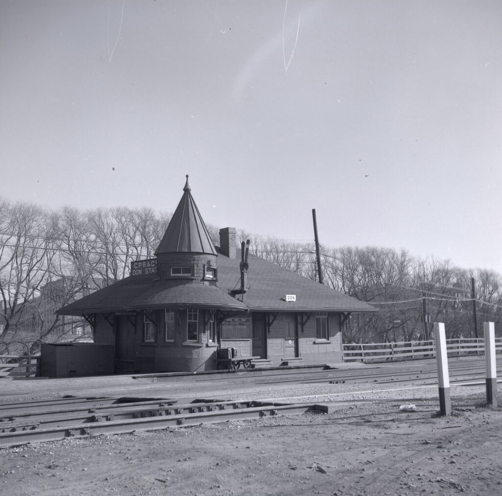 Black and white historical photo of Don Station taken at track level facing the station almost head-on. There is a baggage cart on the platform and the ground around the tracks is bare dirt.