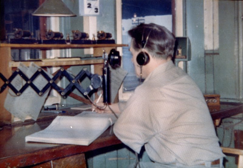 A man sits with his back to us at a desk inside Don Station. He is wearing an over-ear headset and speaking into an old fashioned telephone receiver. In one hand, he holds a pencil over a ledger book.