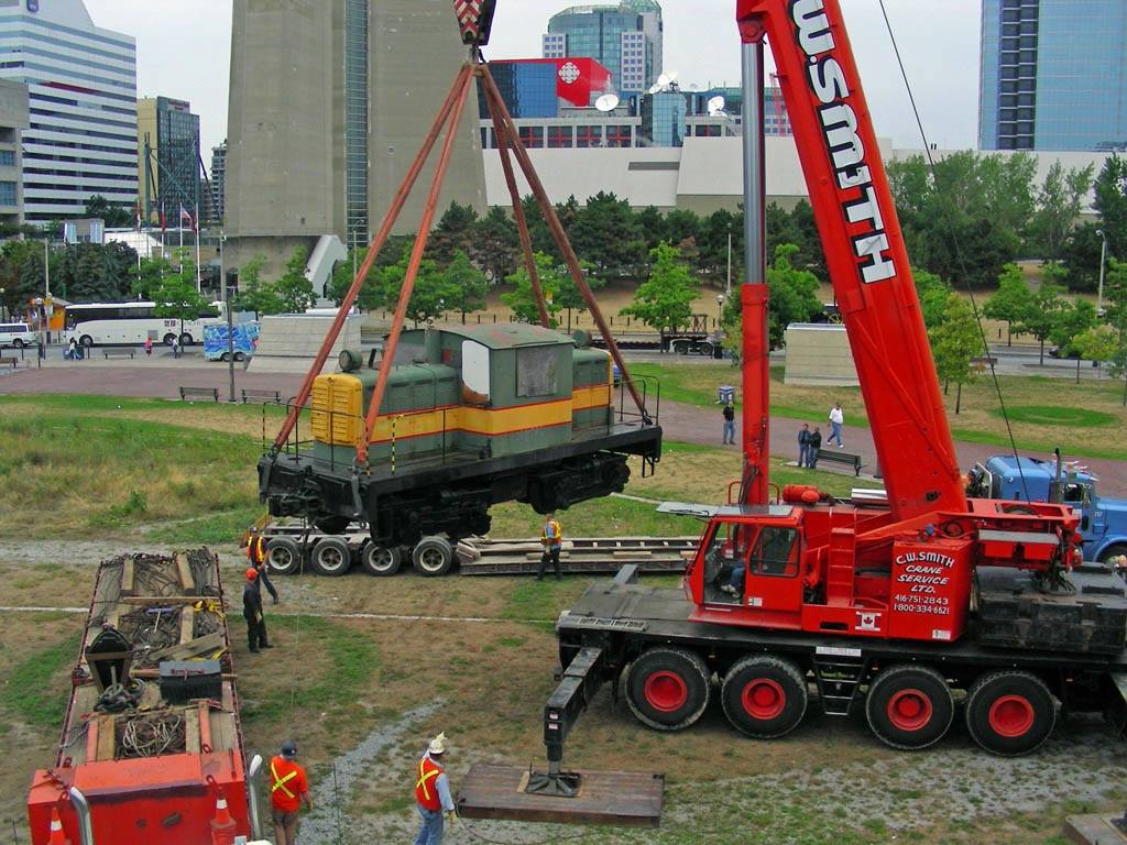 No. 1 is suspended above Roundhouse Park by a massive crane. The locomotive is lifted by thick straps onto a set of tracks on the ground. Workers and the public are gathered around the crane.