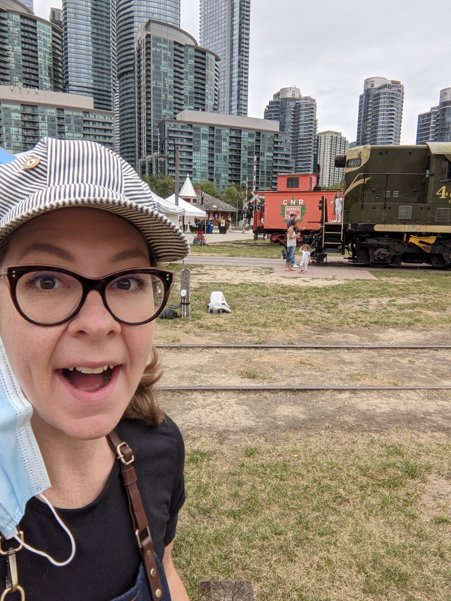 Selfie of Max in Roundhouse Park. She is wearing a striped conductor's cap and several train cars can be seen behind her.