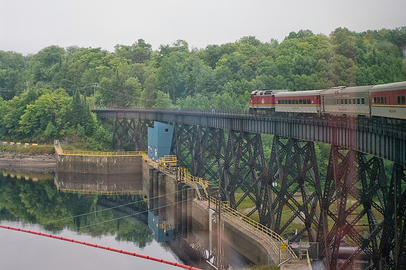 A diesel train hauls passenger cars across a metal bridge. The photo is taken from a passenger car towards the back of the train and a dam can be seen under the bridge.