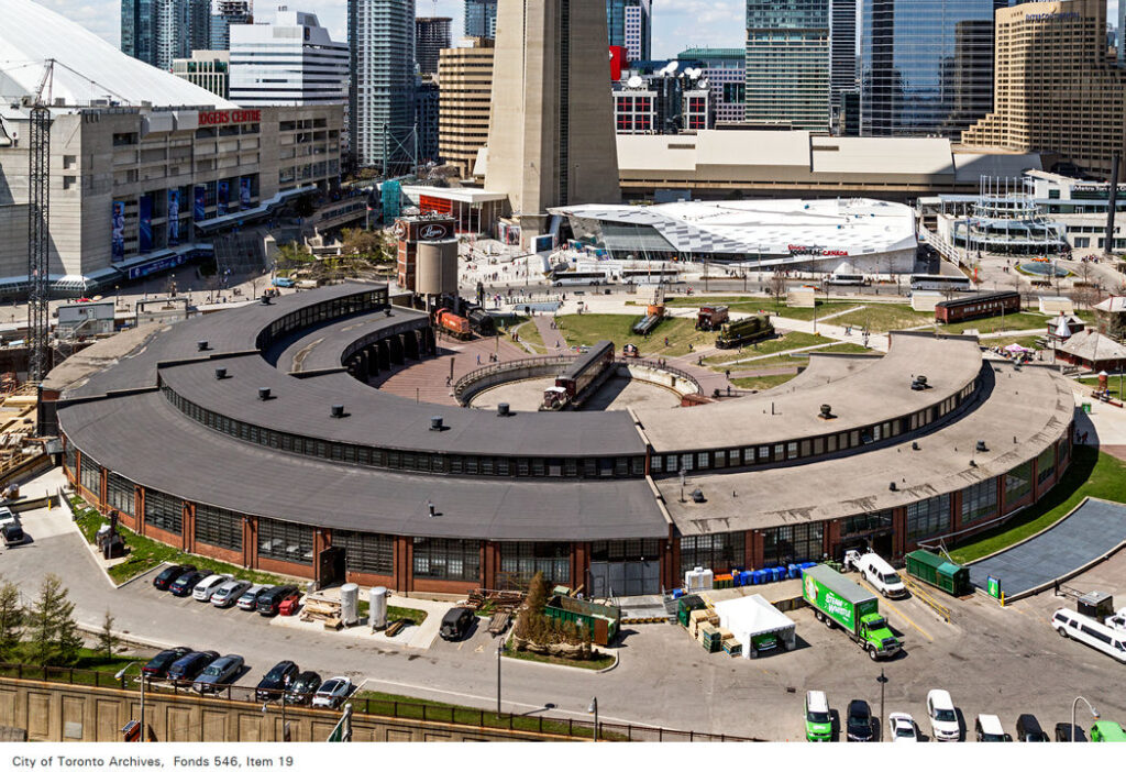 Aerial view of a semi-circle roundhouse building with cars and trucks parked on one side. The base of the CN tower is seen on the other side of the building