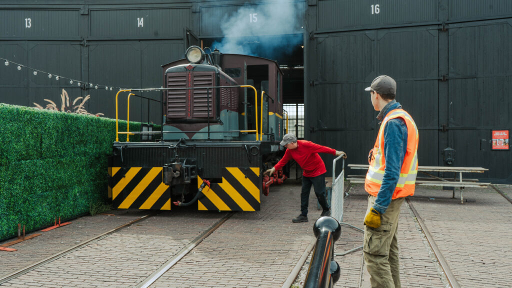 Two volunteers watch a diesel locomotive emerge from the Roundhouse