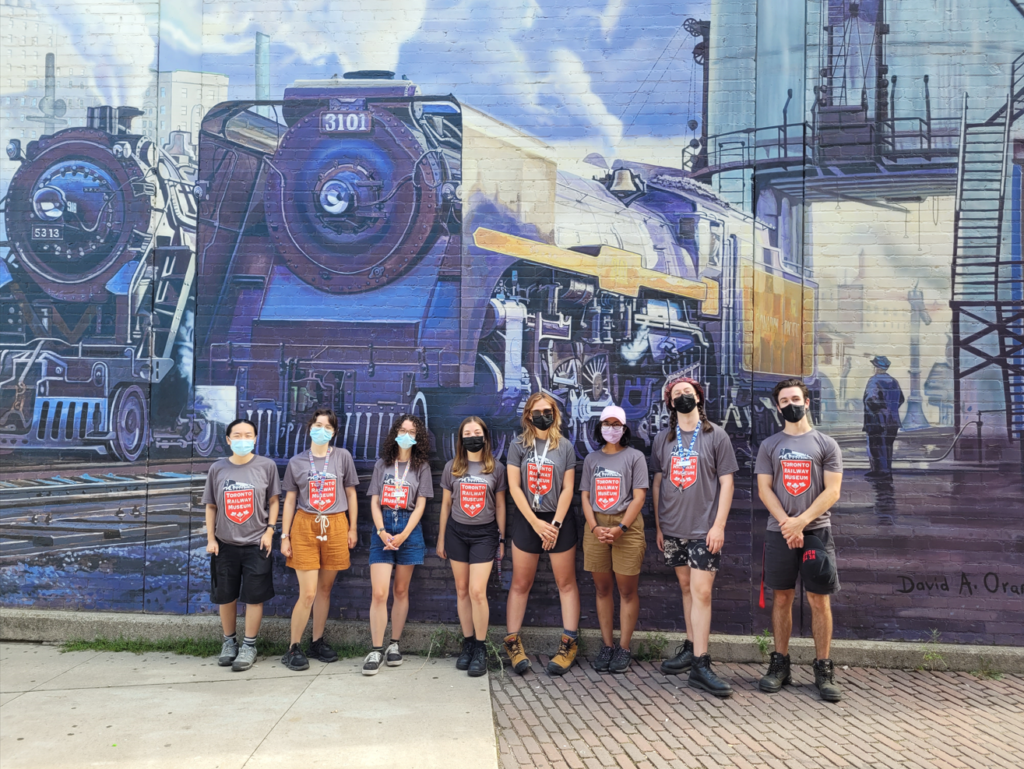 Eight museum staff pose in front of a colourful locomotive mural